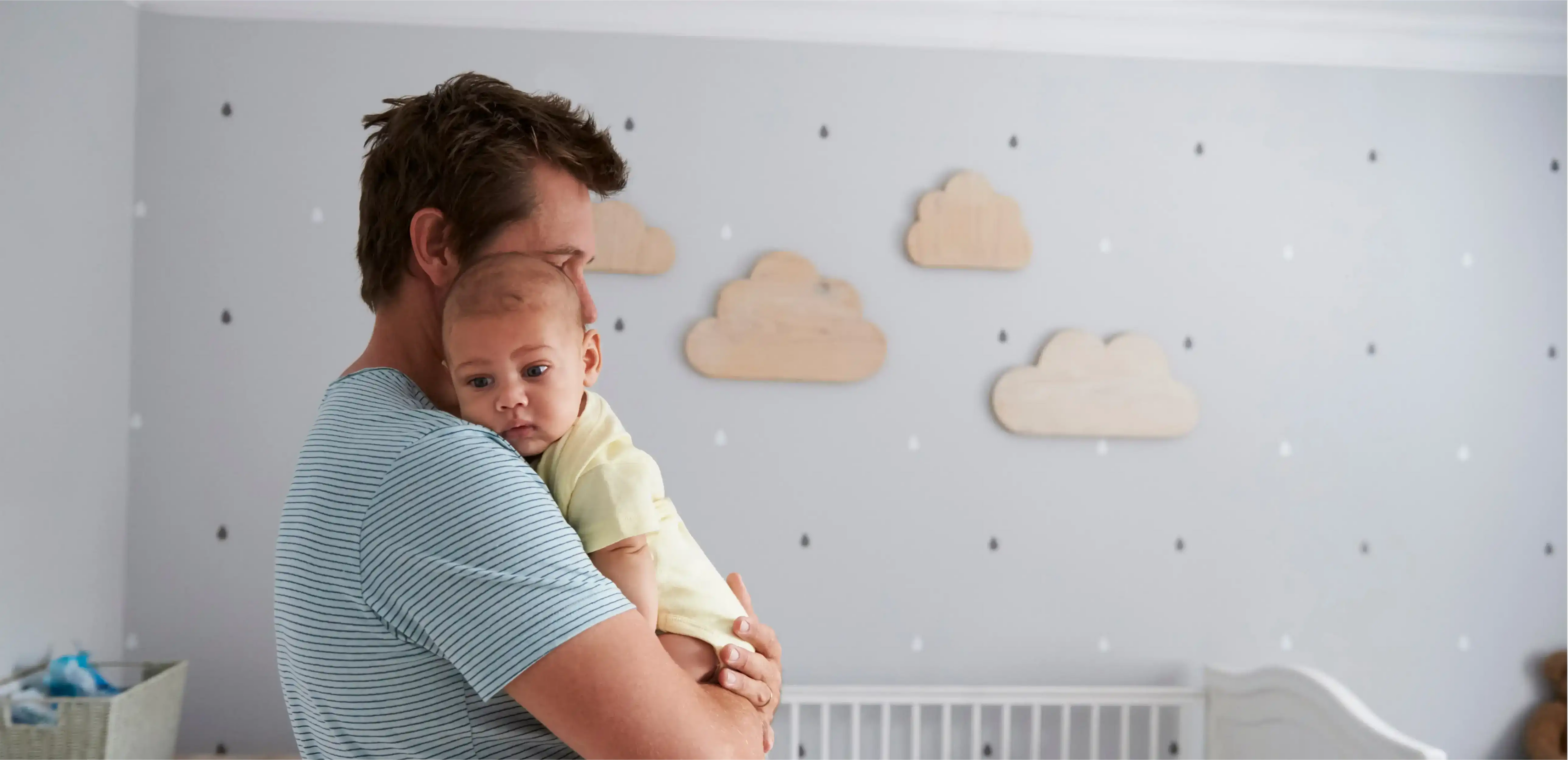 alt= An adult holding a baby in a nursery decorated with cloud-shaped wall decals and a crib in the background, possibly reflecting the joy of new beginnings through services like sperm freezing.